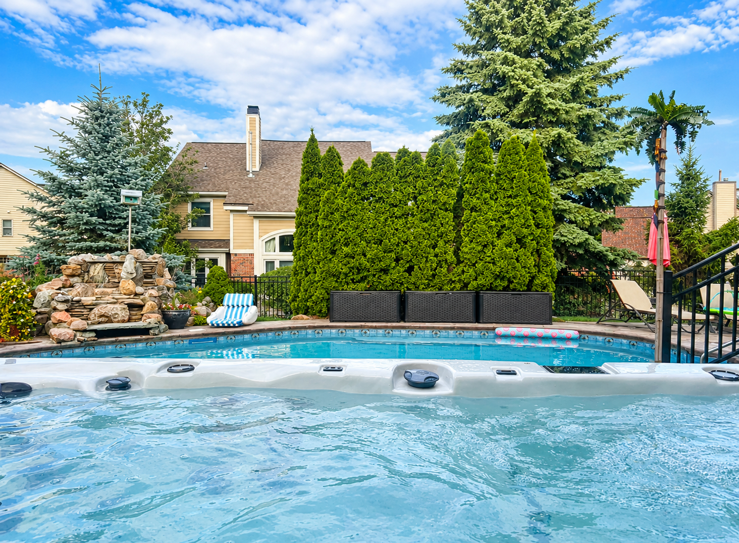 Backyard swimming pool with hot tub, green hedges, rock feature, and patio under a blue sky