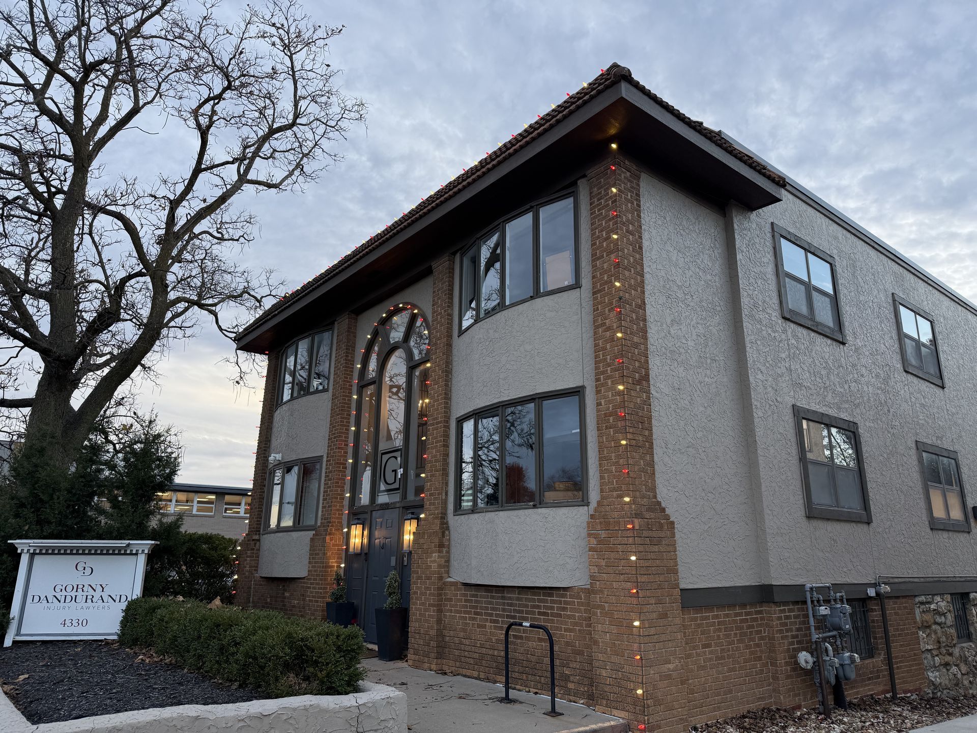 Apartment building with brick accents, arched entrance, and a sign in front. Gray sky, leafless tree.