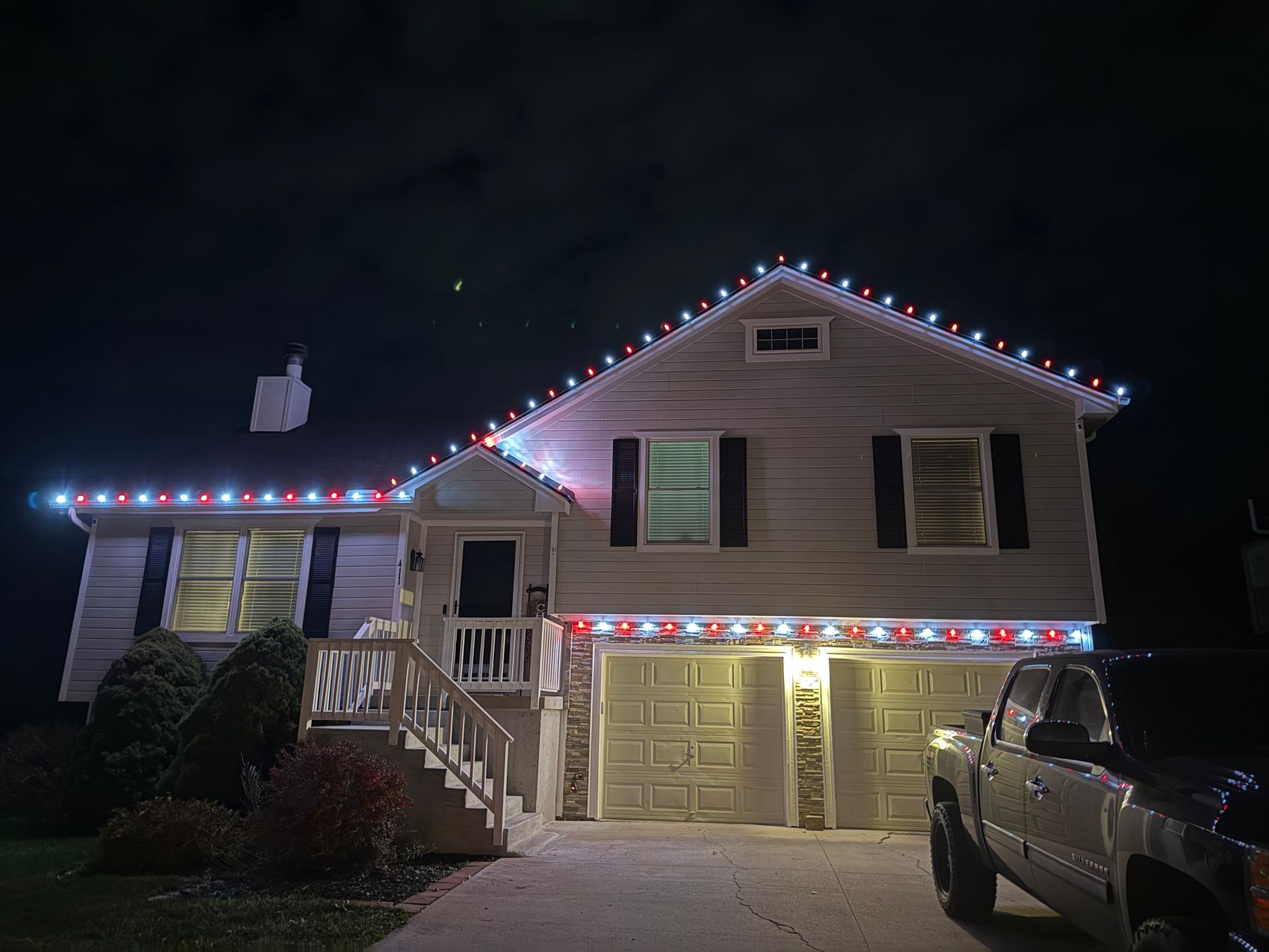 House decorated with red, white, and blue Christmas lights at night, with a truck parked in the driveway.