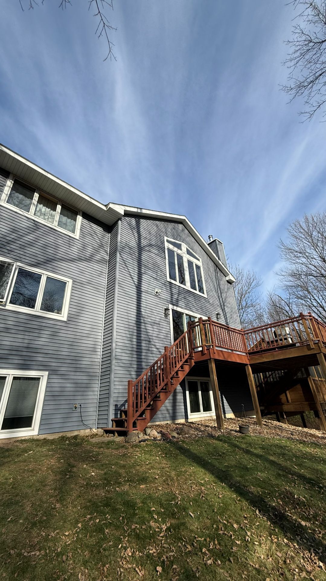 A low-angle view of a grey multi-story house with a wooden deck and staircase under a blue sky with thin clouds.