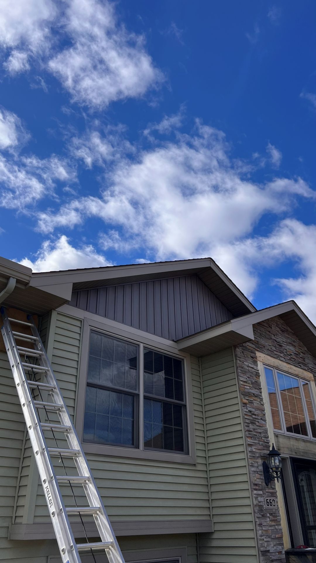 A ladder leans against the side of a house with pale yellow siding, stone veneer, and stained wooden gable trim.