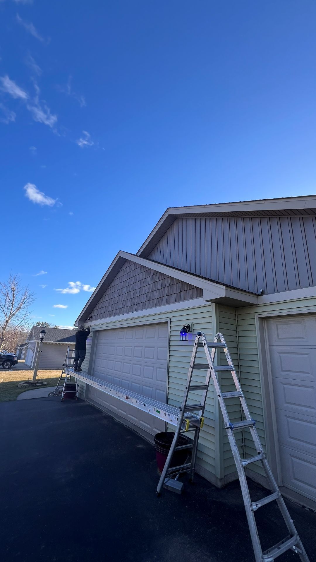 A person stands on a long scaffolding platform attached to ladders, working on the siding of a light-colored garage.