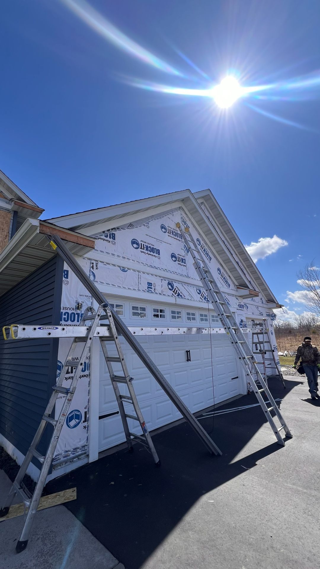 A house under construction with white protective wrap on the garage wall, two metal ladders leaning against it, under sun.