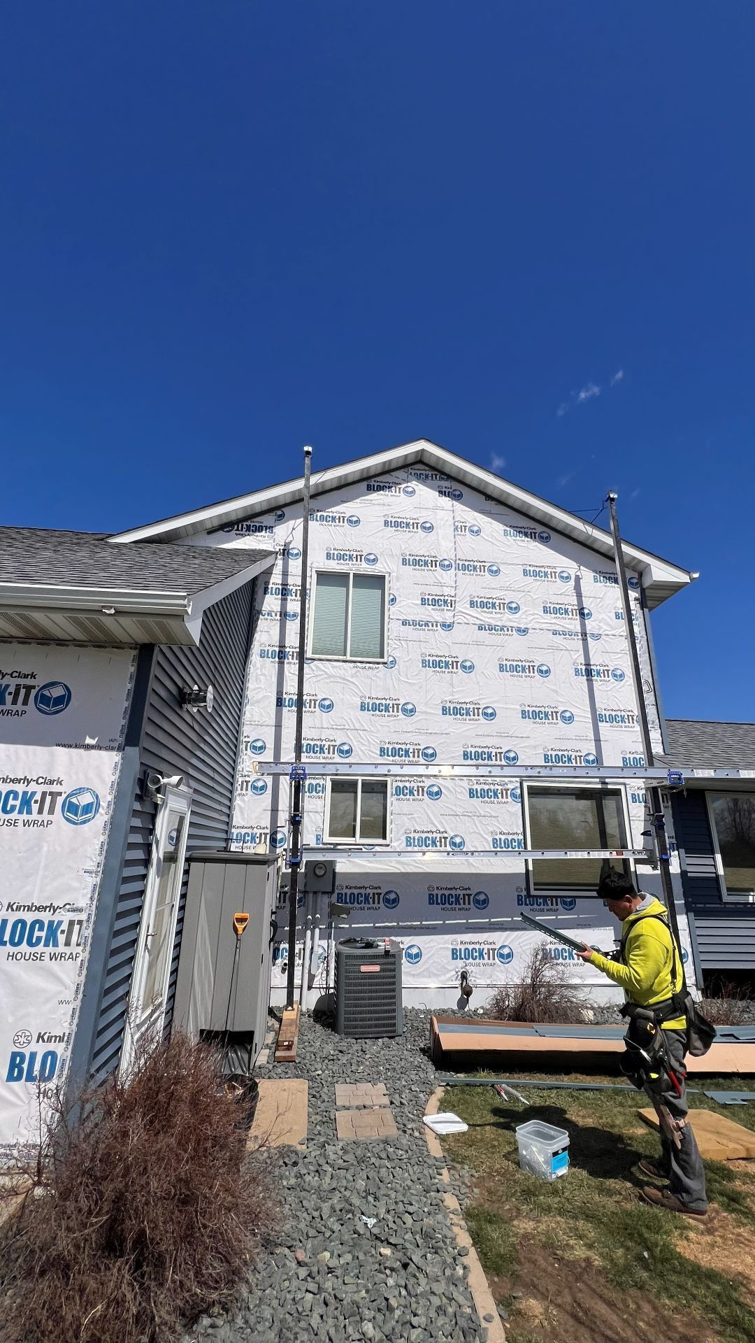 A construction worker in a yellow jacket stands by scaffolding in front of a house covered in white building wrap.