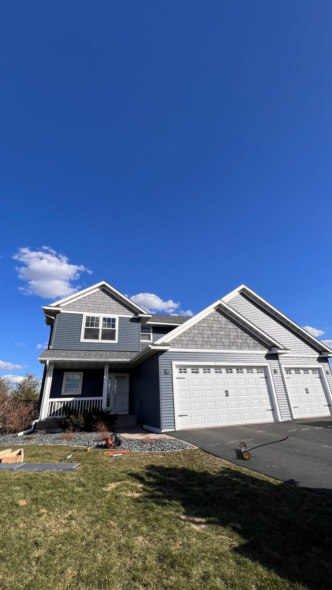 A two-story gray and white suburban house with a multi-car garage under a bright blue sky with scattered clouds.