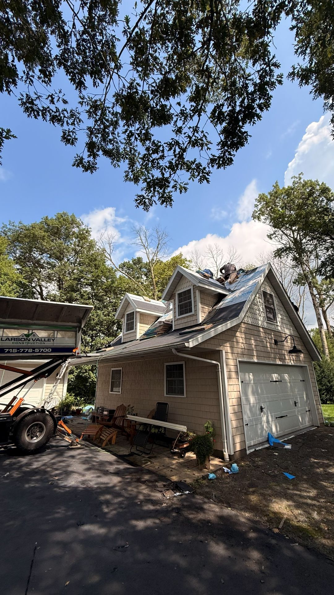 Workers replacing the roof on a light-colored, two-story house with an attached garage, viewed from a sunny driveway.