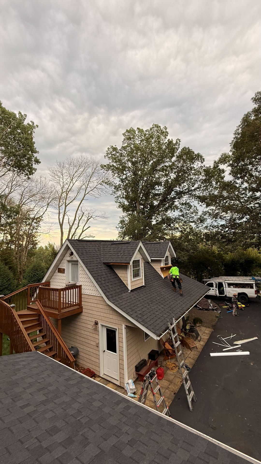 A worker in a bright yellow shirt stands on the shingled roof of a two-story beige building with outdoor stairs.