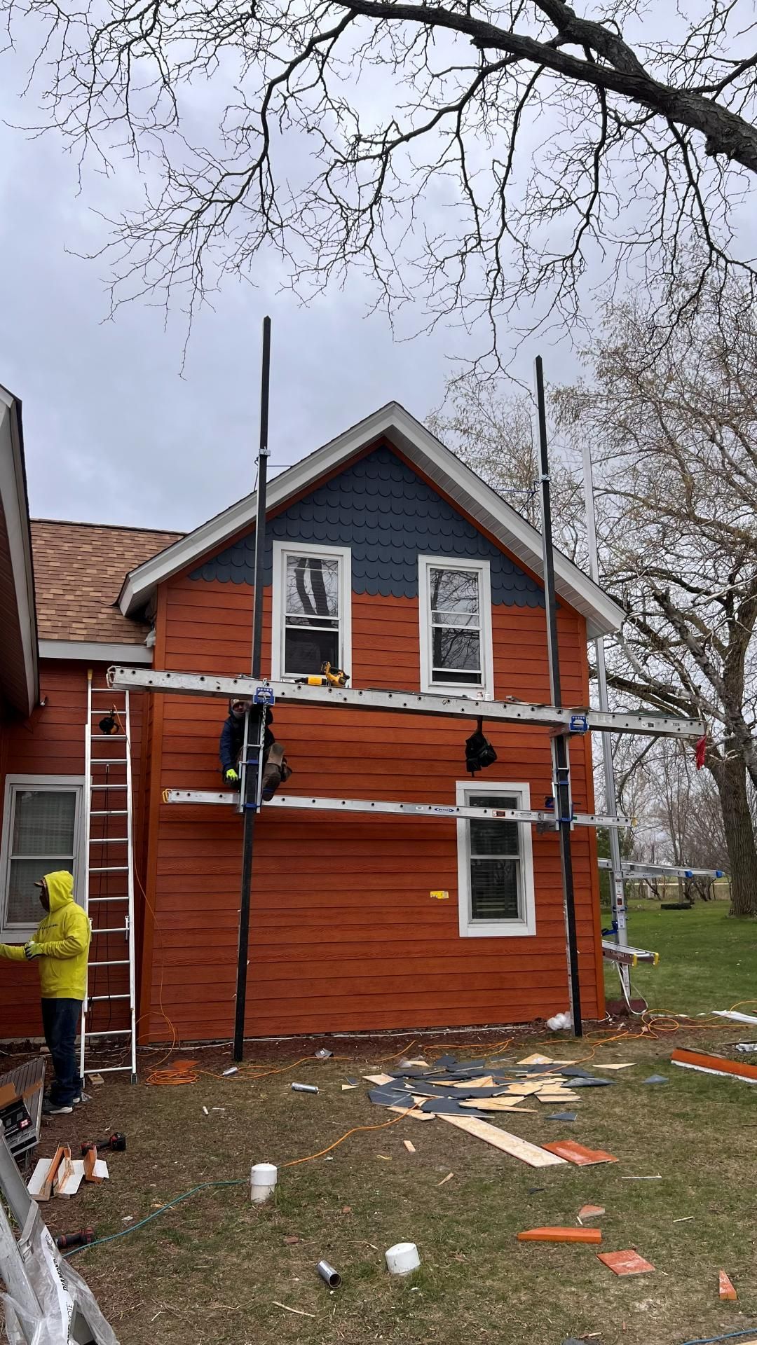 A person in a yellow jacket stands by a ladder next to a house under renovation with scaffolding and new siding.