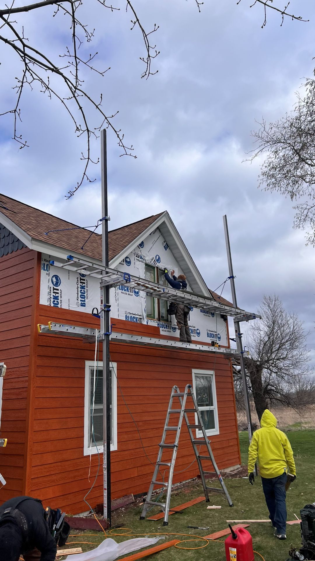 Workers use scaffolding and a ladder to renovate the orange, wooden exterior of a house under a cloudy sky.