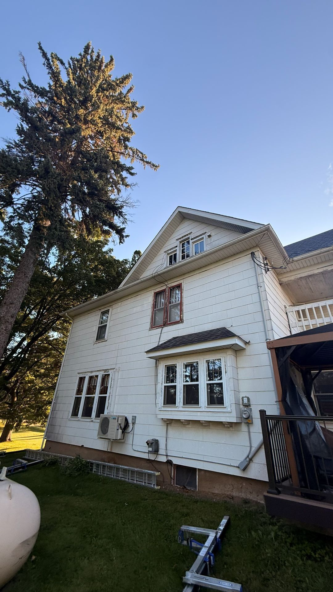A two-story white house with a tall pine tree to its left under a clear blue sky.
