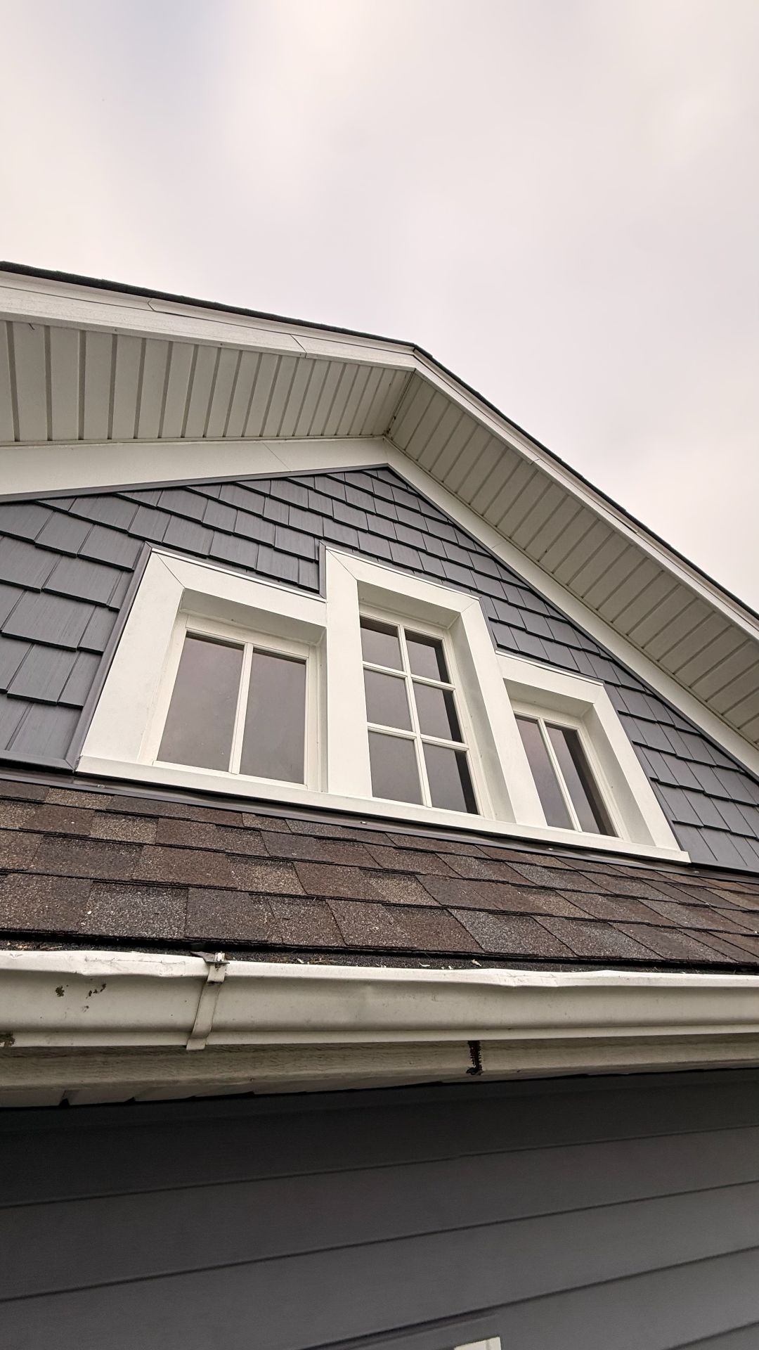 Low-angle view of a house gable featuring blue shingle siding, a dark shingled roof, and three white-framed windows.