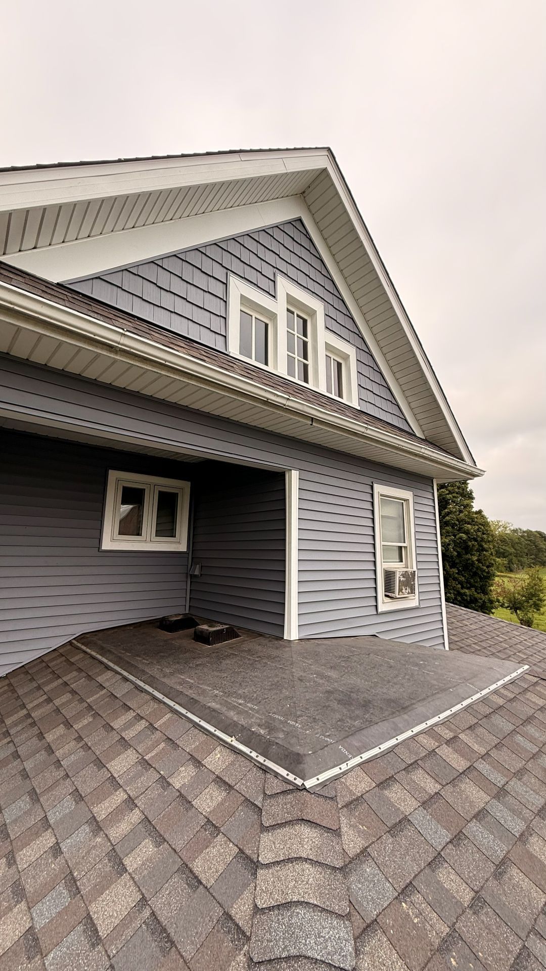 A grey-sided house with a shingled roof featuring a low-slope roof section and white window frames under a cloudy sky.