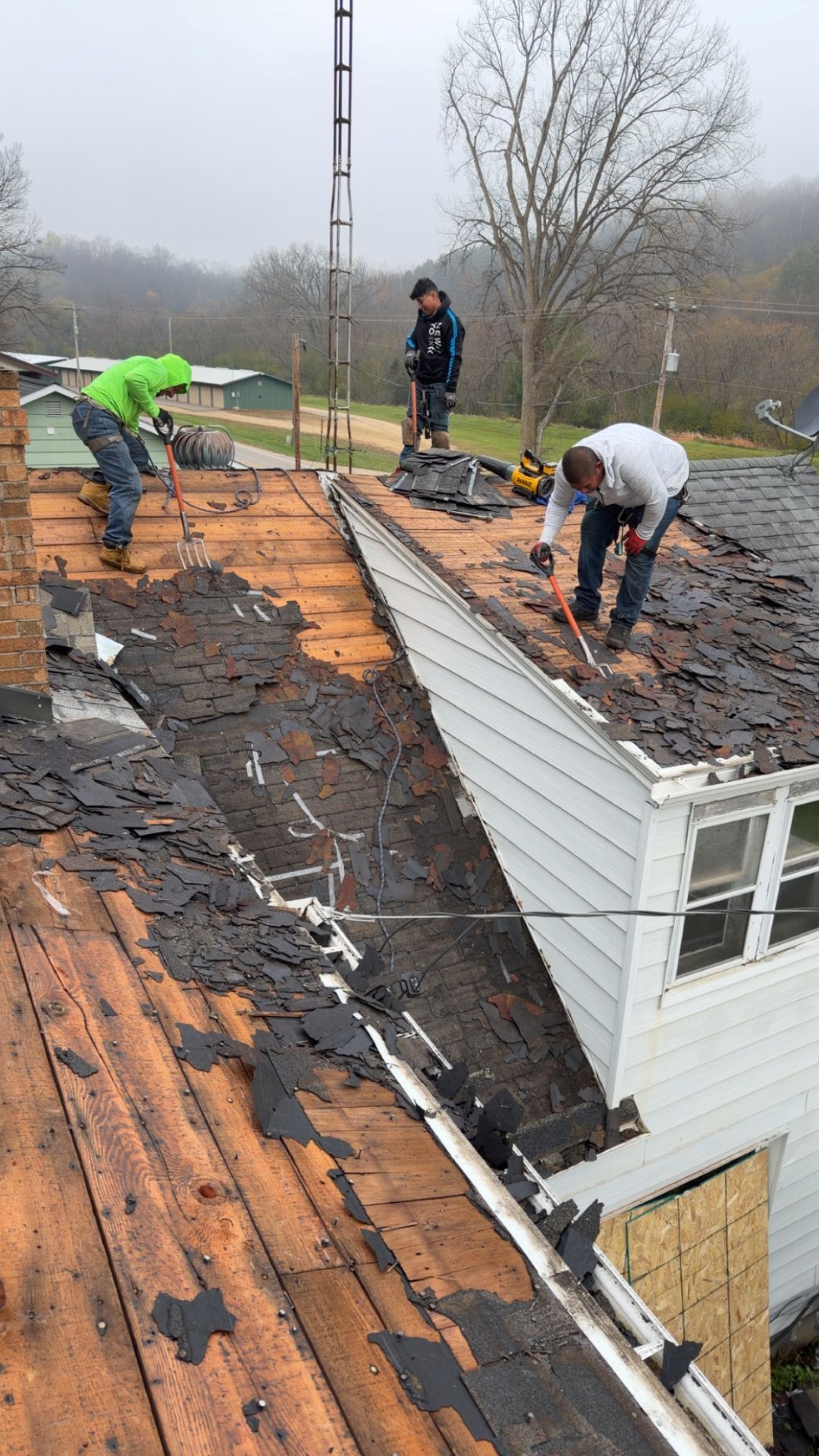 Three workers wearing safety gear are removing old asphalt shingles from a residential roof.