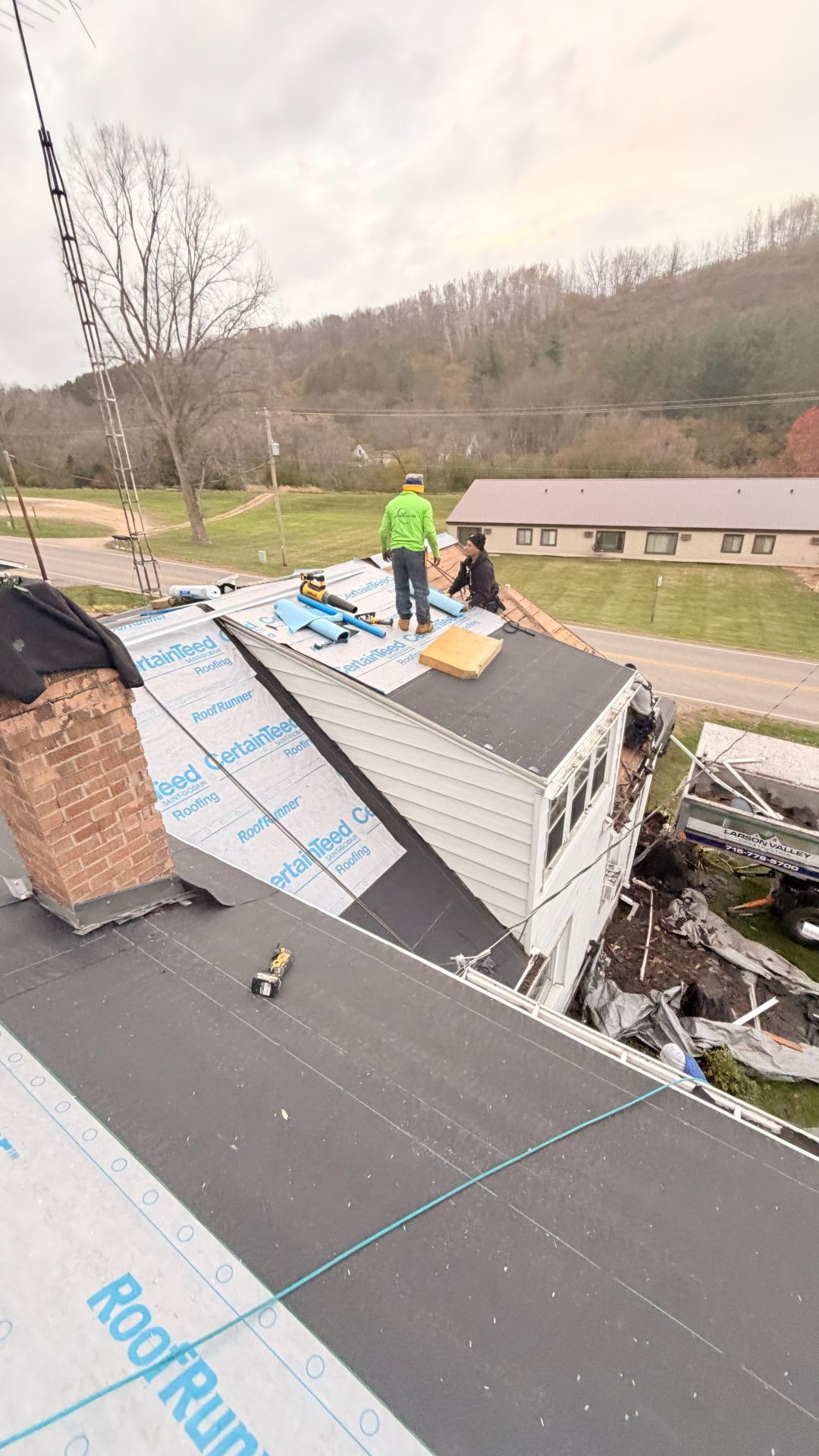Two workers install new roofing material and underlayment on a multi-level residential building on a cloudy day.