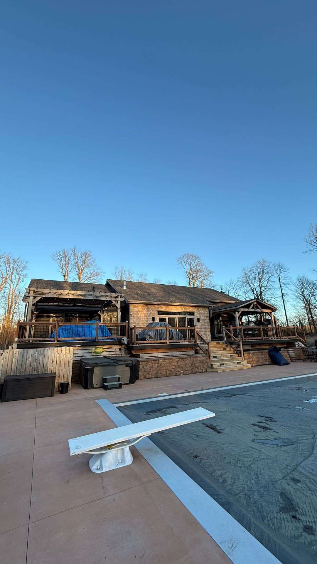 A backyard swimming pool with a diving board faces a house with large decks and stone exterior walls under a clear sky.