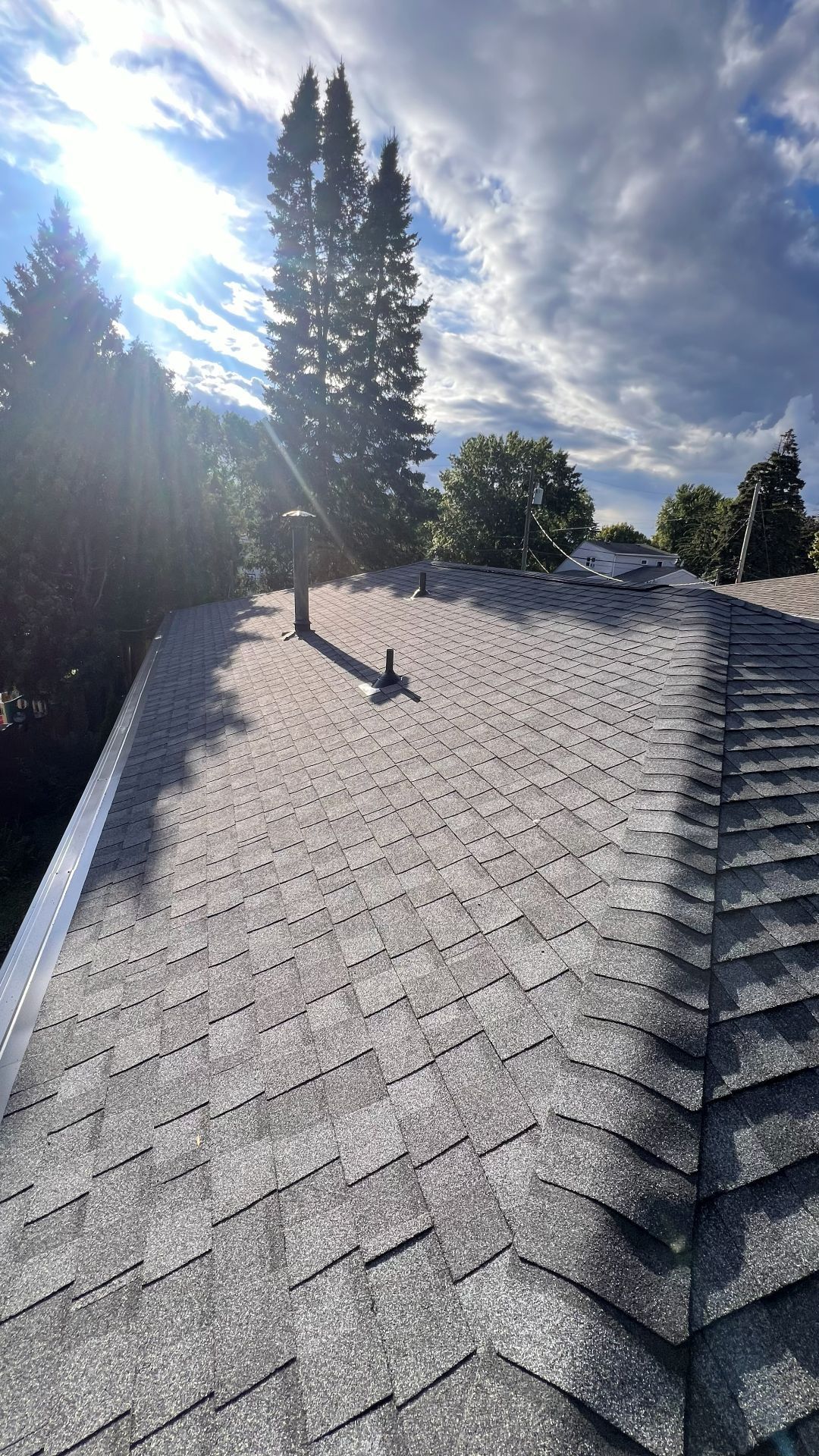 A view looking up a sloped, gray shingled roof with two vent pipes under a bright sun and blue, cloudy sky.