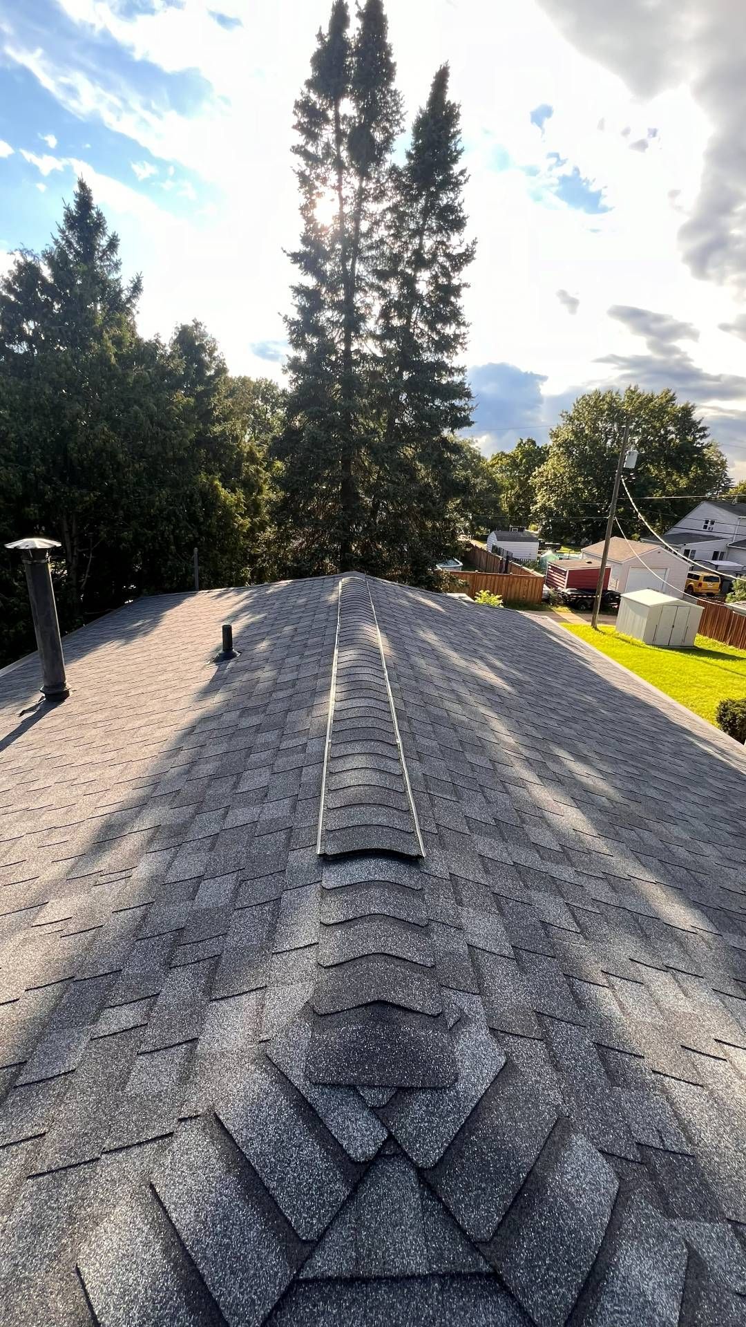 A view looking down the shingled ridge of a residential roof toward tall pine trees under a cloudy sky.