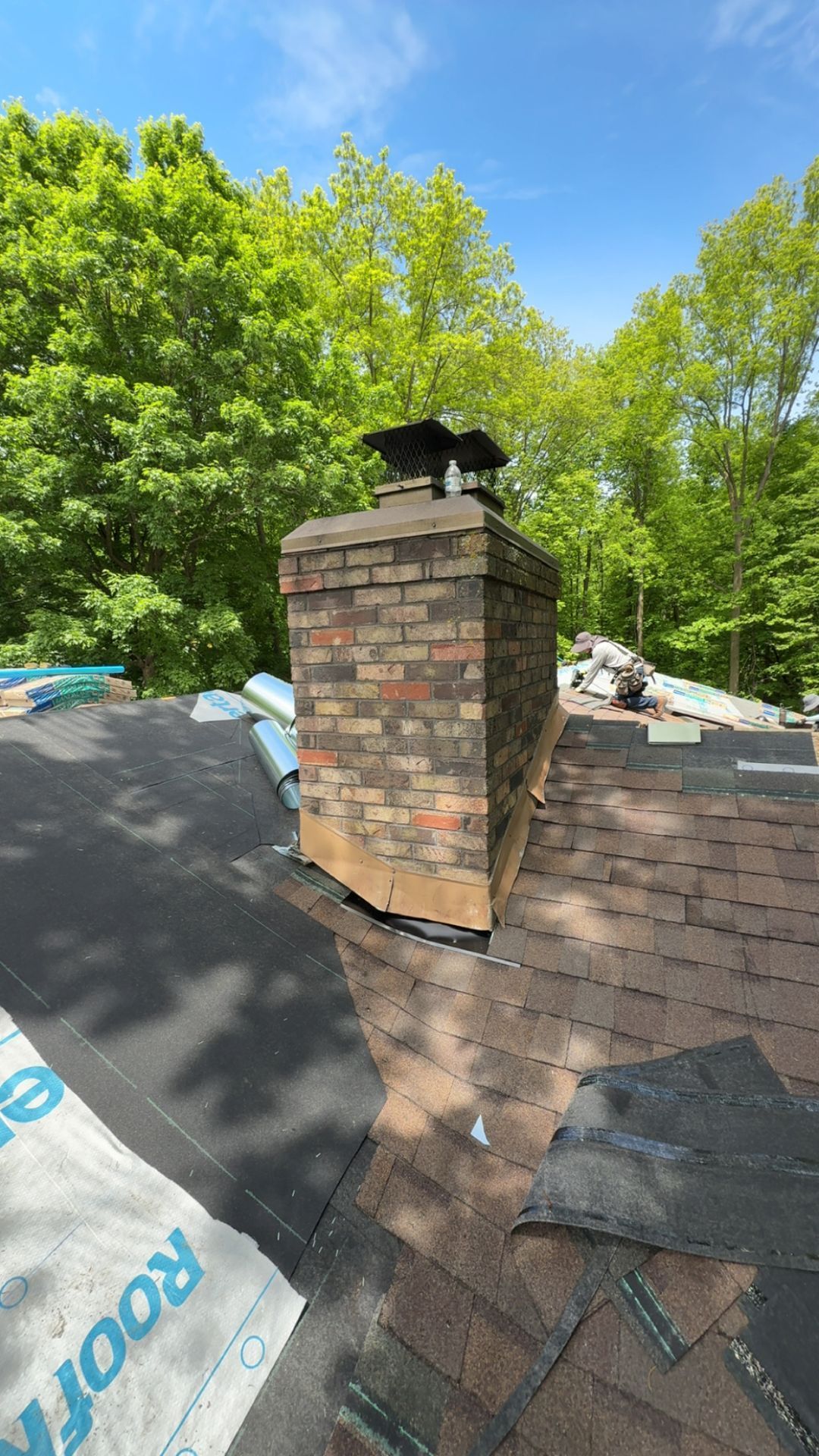 A brick chimney on a house roof under construction, featuring exposed roofing felt and new shingles against a treed backdrop.