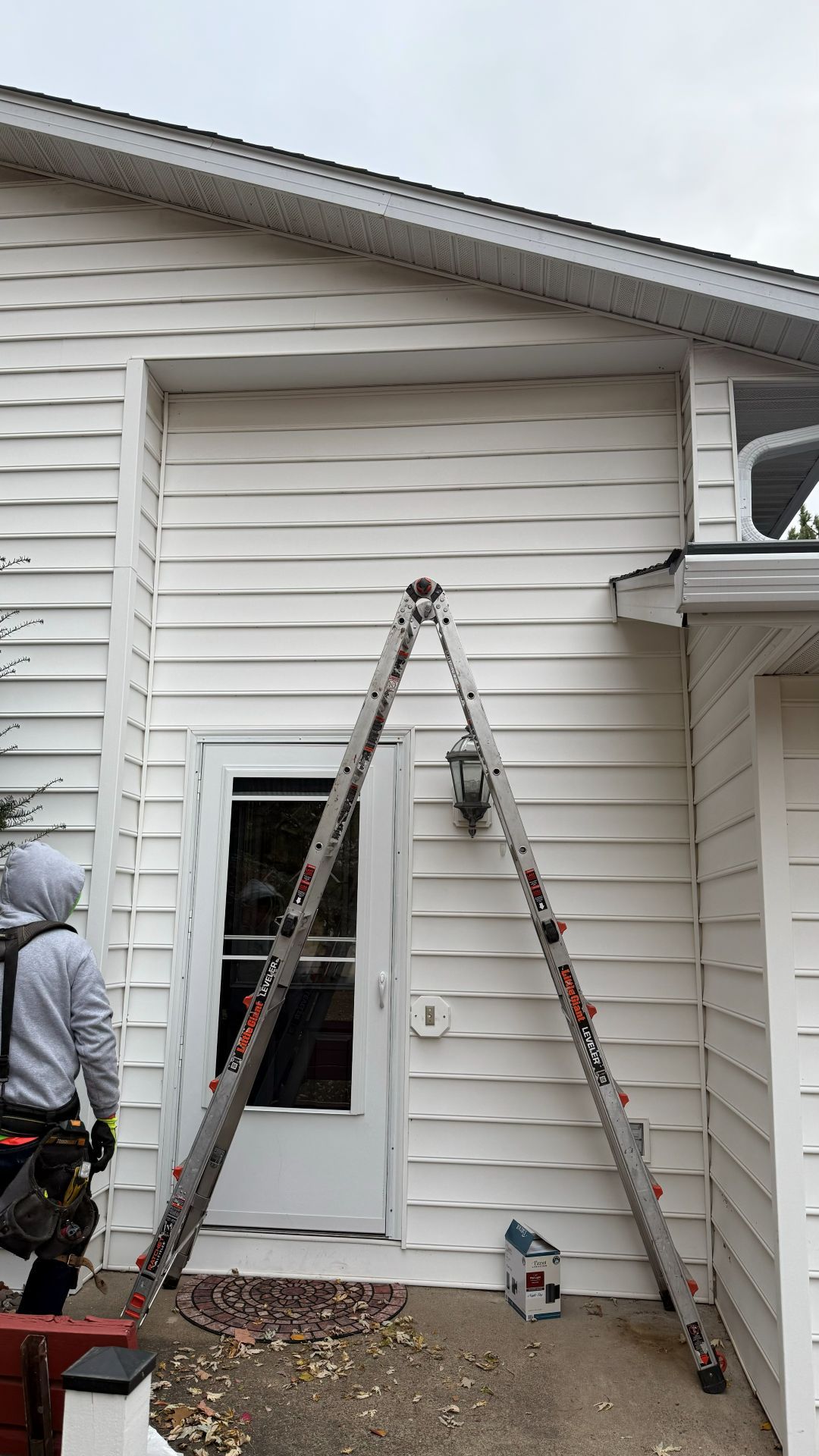 A person in work gear stands near a tall A-frame ladder positioned against the white vinyl siding of a house exterior.