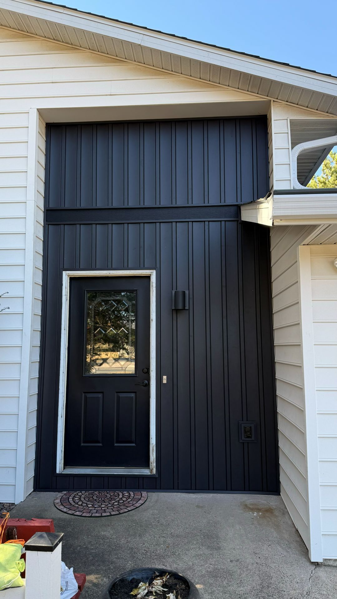 Black vertical siding surrounds a front door on a white house, with a small welcome mat on the concrete porch.
