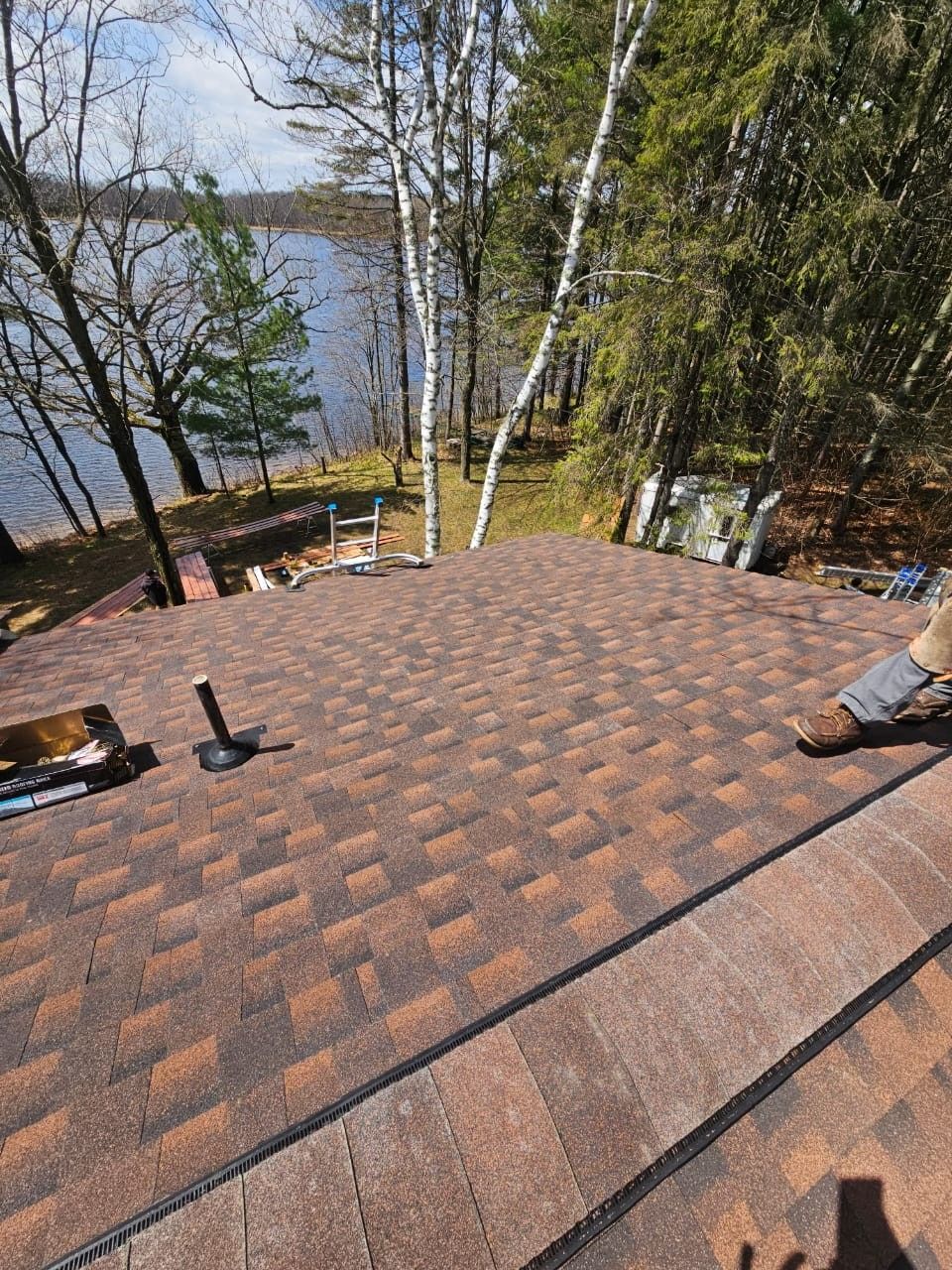 A high-angle view of a newly shingled brown roof overlooking a lake and trees, with a plumbing vent visible.