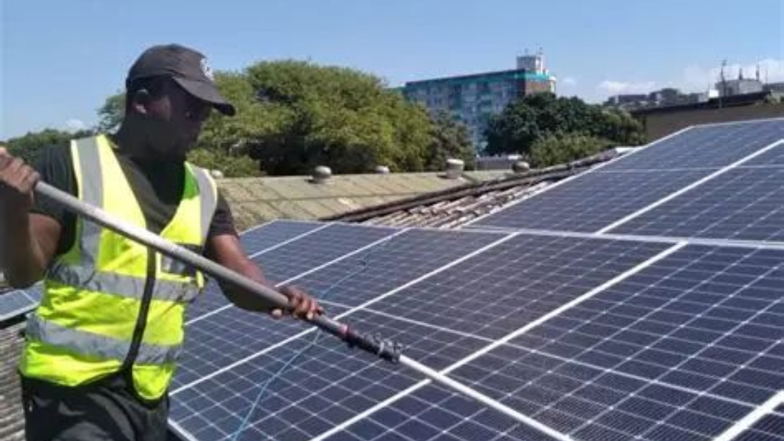 Man cleaning solar panels on a roof with a long-handled brush, wearing a safety vest. Blue sky in the background.