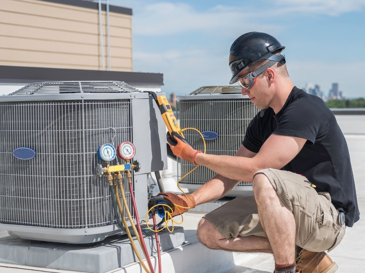 Technician crouches by an outdoor HVAC unit, using a yellow tool to service gauges and hoses.