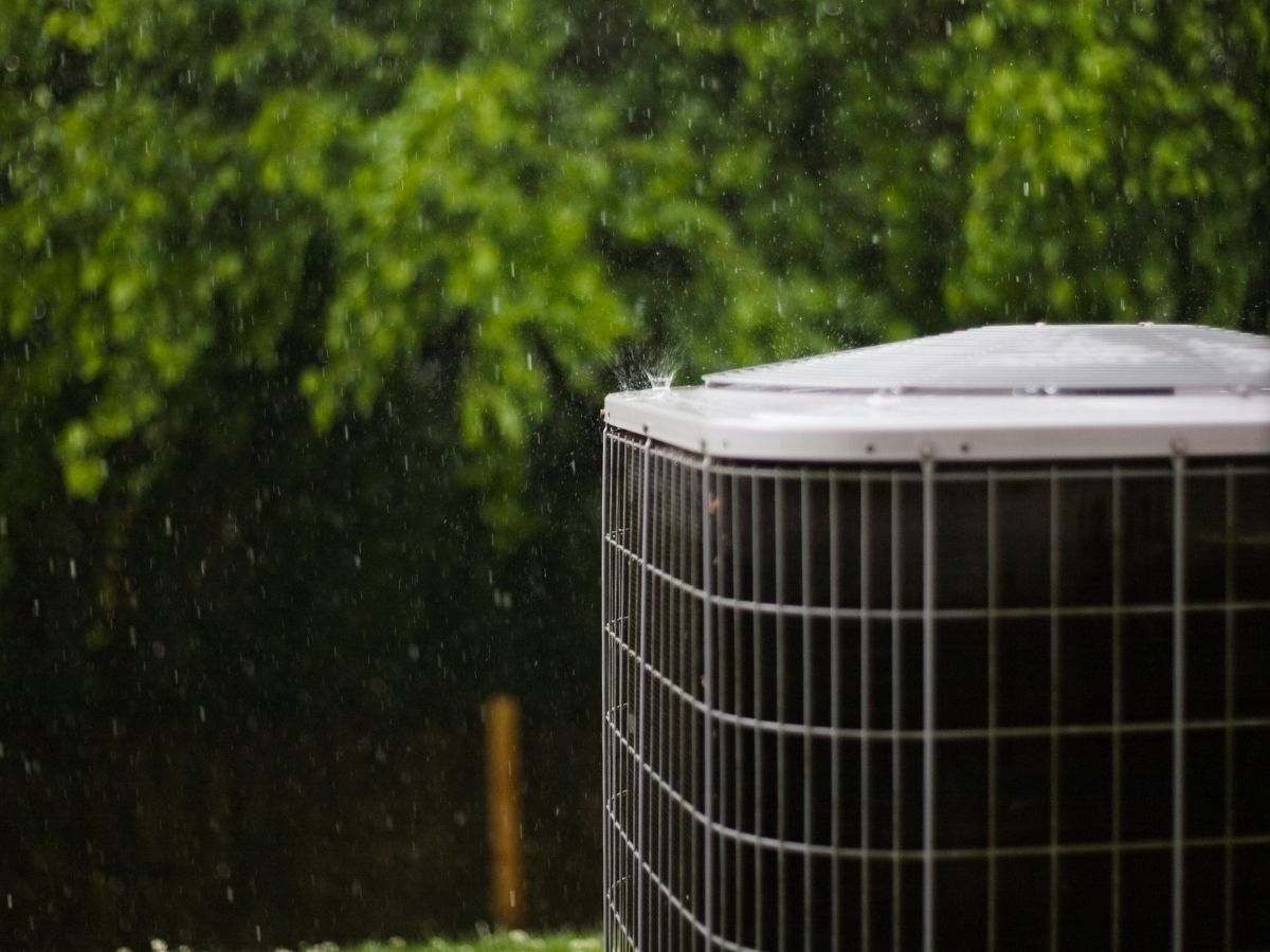 Outdoor air conditioner unit in rain beside green foliage