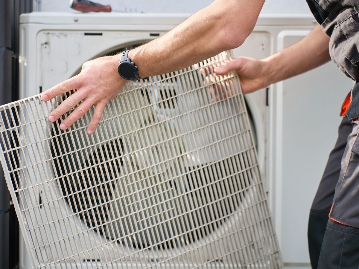 Hands removing a large white fan grille from an outdoor air conditioner unit