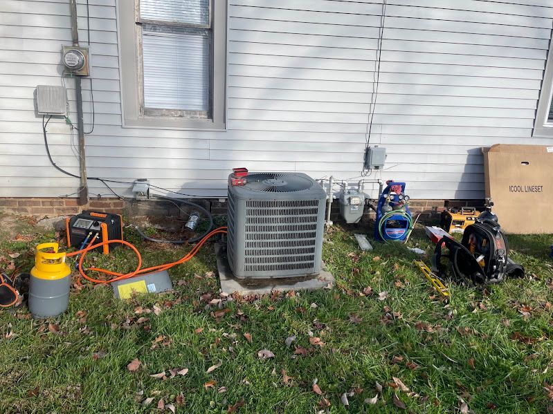 Outdoor HVAC equipment and tools along a house wall, with ducts, cords, and a covered unit in a yard.