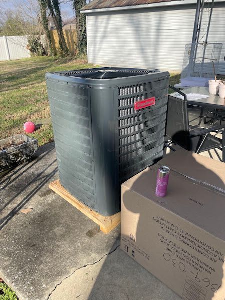 Outdoor HVAC unit on a wooden platform beside a garage, with a cardboard box and purple can in the foreground.