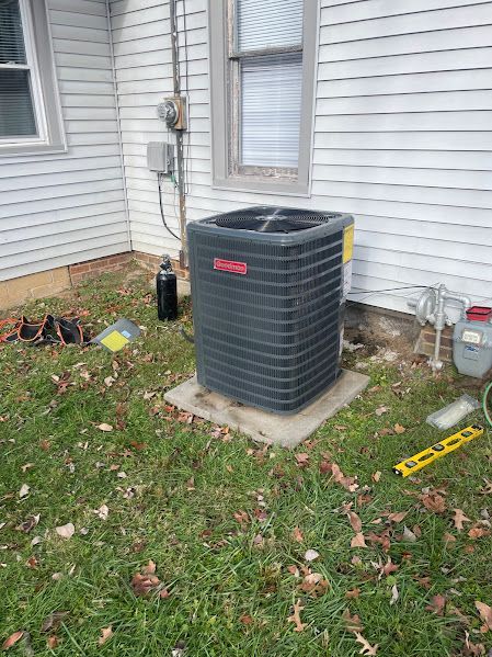 Outdoor air conditioner unit beside a white house, with tools and scattered items on the lawn.