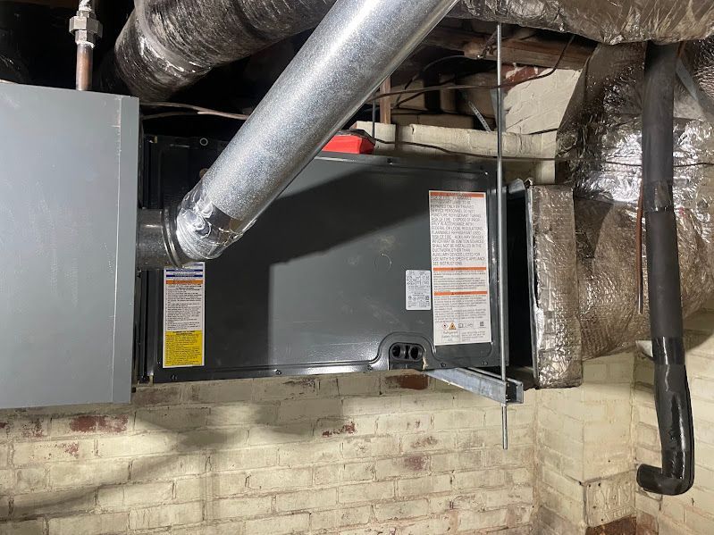 Furnace in a basement with metal ductwork, gray panels, and exposed brick wall.