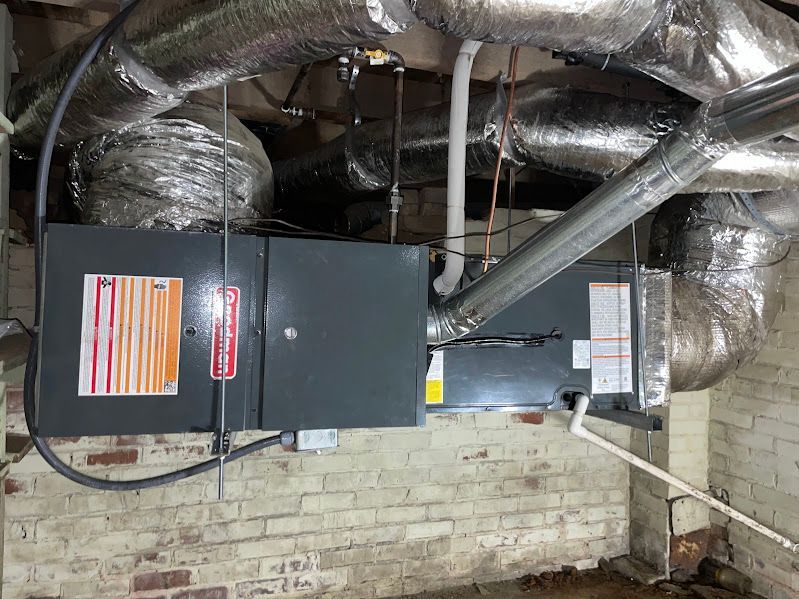 Ductwork and two HVAC units mounted in a basement, with insulated silver ducts and a brick wall backdrop