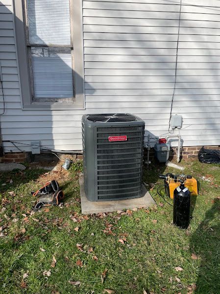 Outdoor air conditioner unit beside a house, with a trash bin and yard items on grass