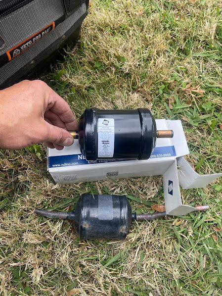 Hand holding a black cylindrical part over a box on grass, with another similar part beside it.