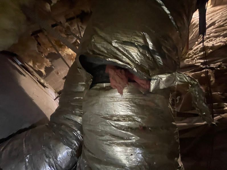Torn silver ductwork in an attic, with exposed insulation and a damaged vent opening.