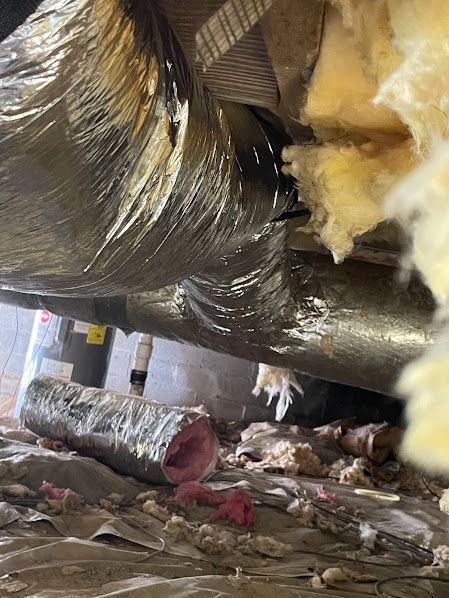 Insulated ductwork under a building, with debris and exposed pipes on the floor beneath it