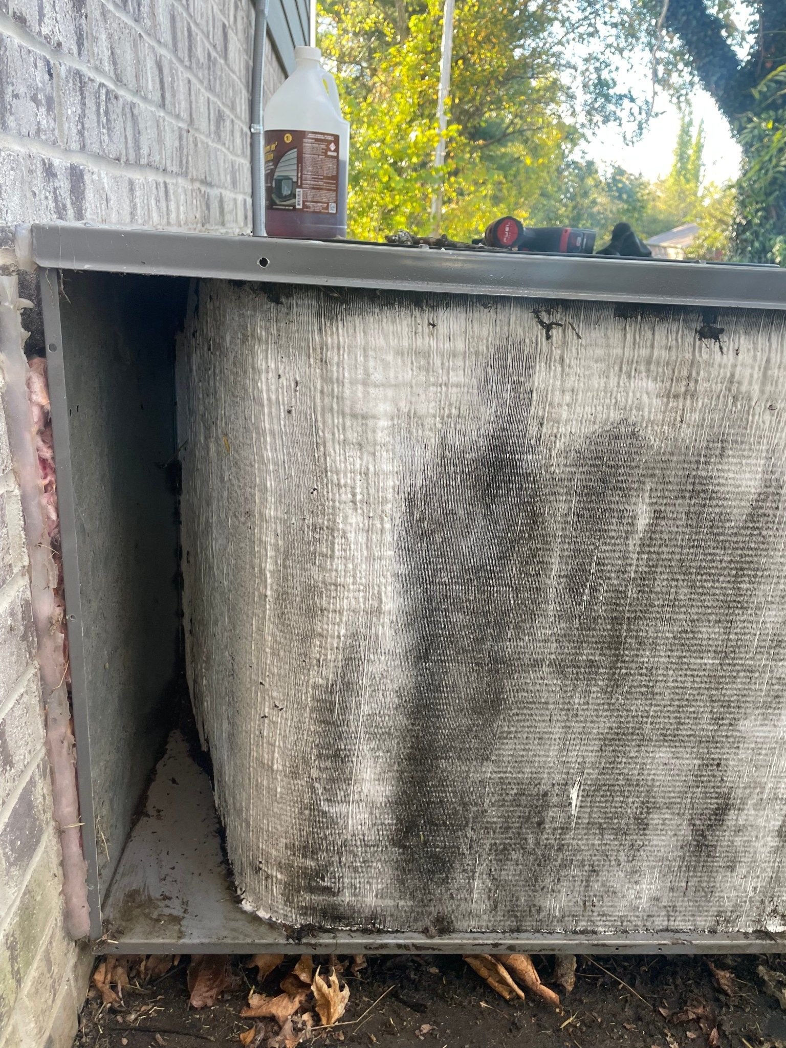 Exterior AC unit beside a brick wall, with a bottle sitting on top and leaves below