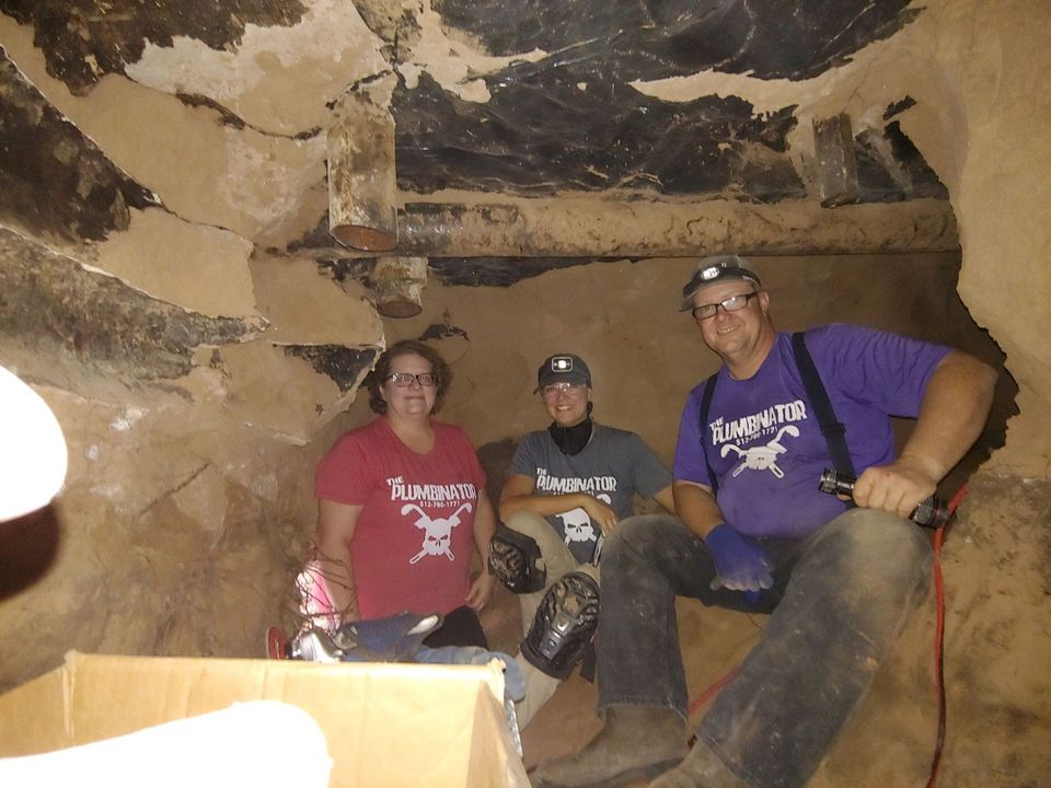 Three people wearing helmets sit inside a rocky cave passage, lit by flash photography.