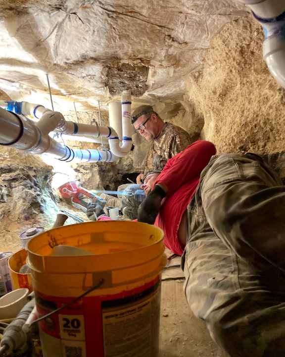 Workers crouch in a cramped underground tunnel beside pipes and buckets.