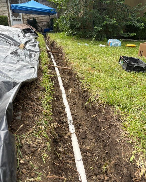 Backyard trench with a white PVC pipe laid in dark soil beside grass and a tarp-covered area