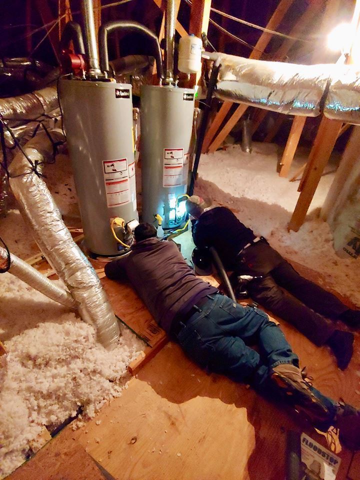 Two workers crawl under a water heater in a snowy crawlspace, inspecting plumbing under a house.