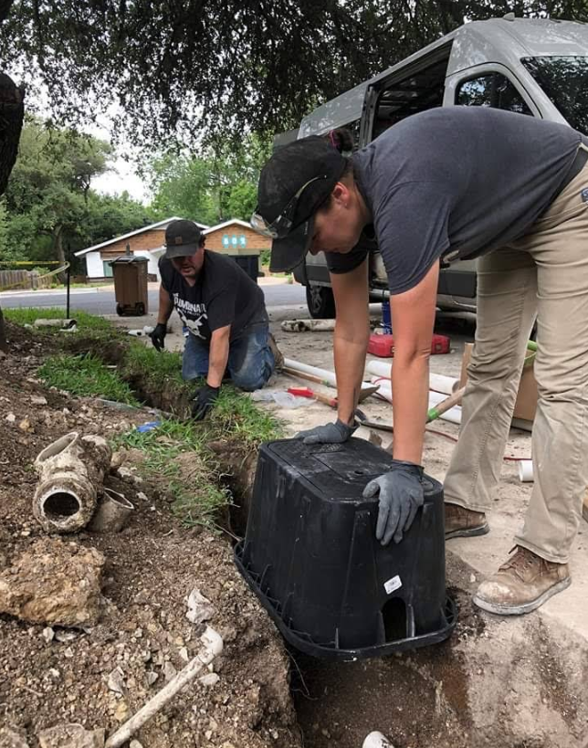 Two workers lift a black tub from a muddy roadside trench near a white van.