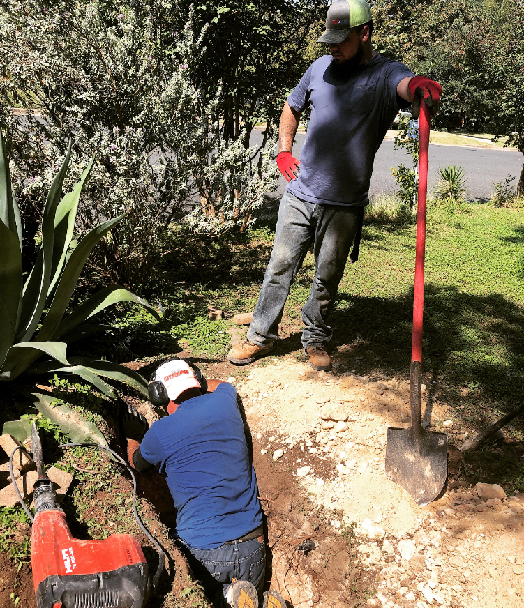 Two workers repairing a dirt path, one digging with a shovel and one standing nearby with a tool.