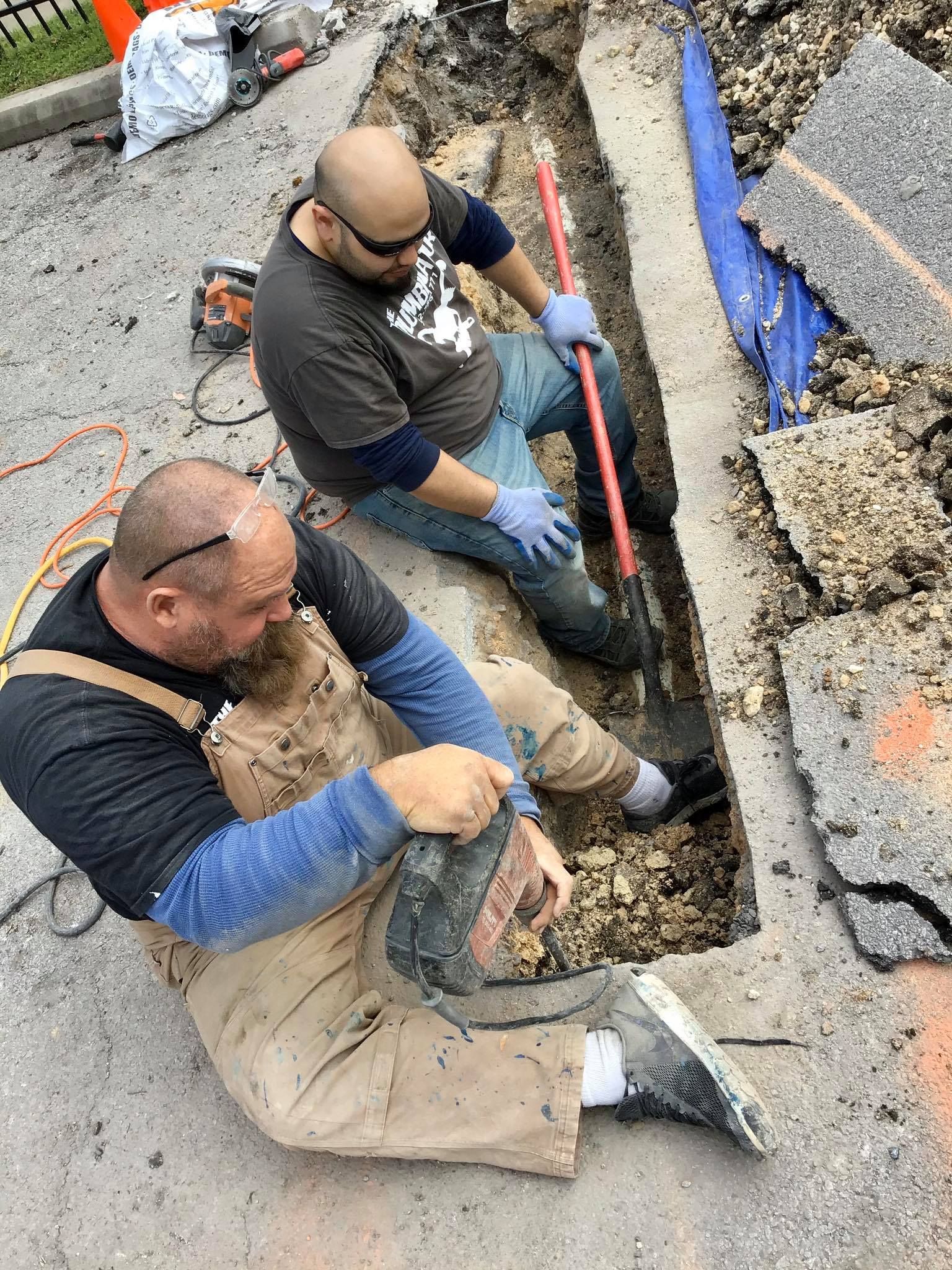 Two workers repairing a broken road trench with tools and blue pipe in a gravel street