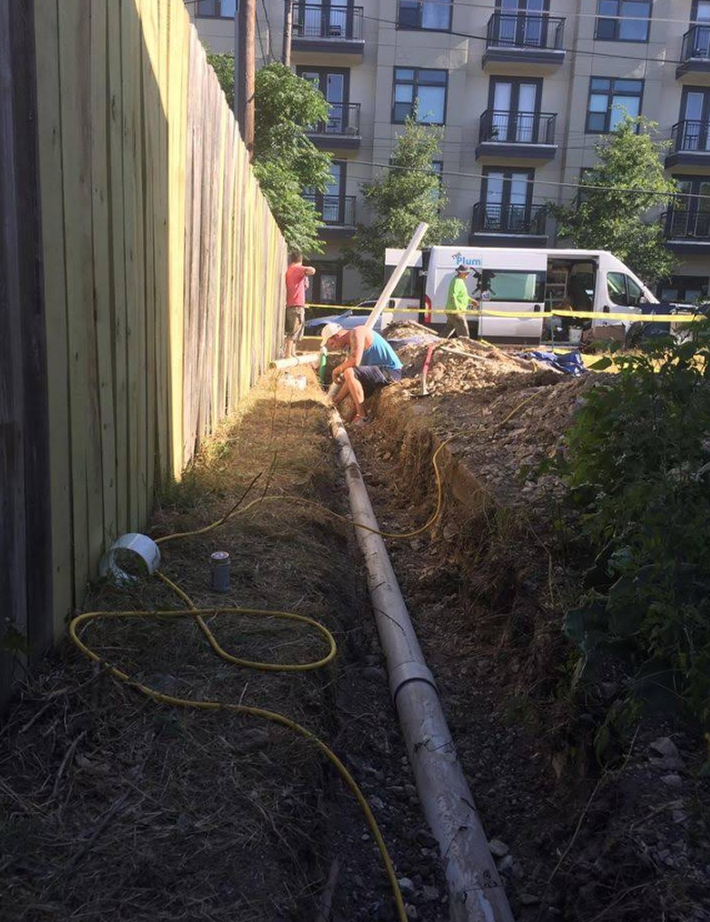 Construction trench with workers laying a long pipe beside a fence and apartment buildings