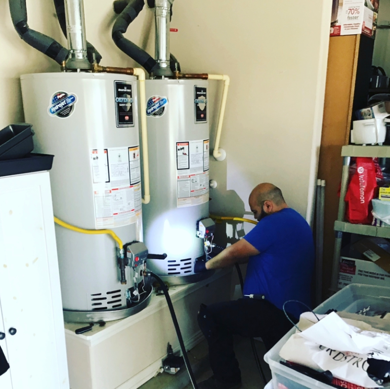 Technician kneeling beside two white water heaters, working on piping in a utility room.