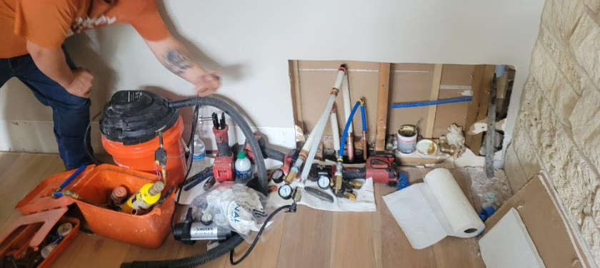 Technician working beside plumbing tools and supplies in a small unfinished room under a sink area