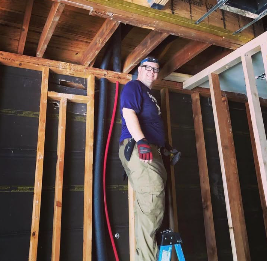 Worker standing on a blue stool inside a framed construction site, wearing a hard hat and gloves.